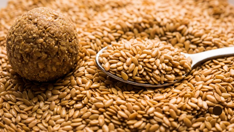 Seeds in Hindi: Names, Benefits & Best Season 10 Golden-brown flaxseeds on a spoon next to a traditional winter alsi laddoo sweet, showcasing alsi ke beej in Hindi.